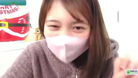 Video of a young Asian woman with long brown hair, wearing a pink mask, grey hoodie, and looking directly at the camera. Background includes a red and white Santa hat and a plush toy.