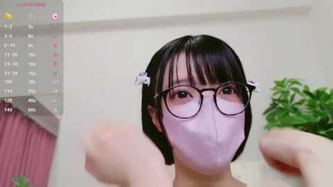 Media: Video of an East Asian woman with short black hair, wearing round glasses, a pink face mask, and a light pink top. She is indoors, with a calendar, a plant, and a pink curtain visible in the background.