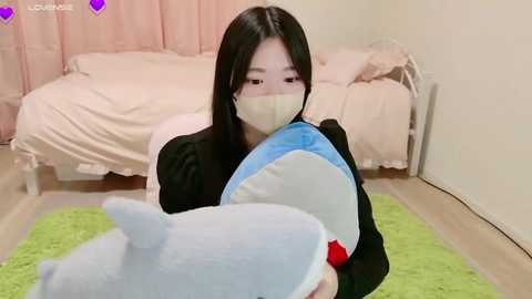 Media: Video of an Asian woman with long black hair, wearing a mask, holding a plush shark in a pastel-colored bedroom with light wood floors and beige curtains.