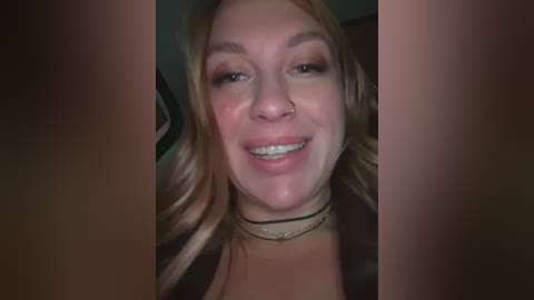 Media: Video of a smiling young woman with light skin and long, wavy, blonde hair, wearing a black choker necklace, and minimal makeup. The background is dark and indistinct.