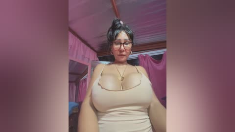 Media: Video of a curvy, light-skinned woman with glasses, black hair tied up, wearing a tight beige tank top that accentuates her large breasts. She stands in a dimly lit room with pink curtains and a wooden ceiling.