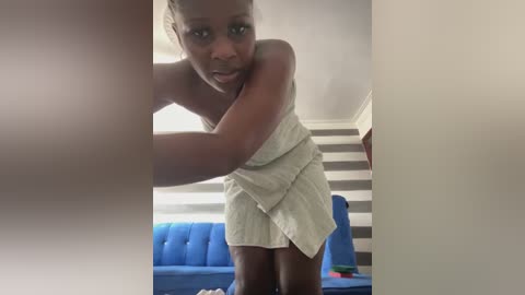 Media: Video of a dark-skinned woman with a towel wrapped around her body, standing in a room with a blue couch and striped wall.