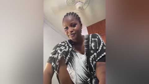 Media: A video of a confident Black woman with braided hair, wearing a black and white patterned blouse, standing in a room with a chandelier and wooden door.