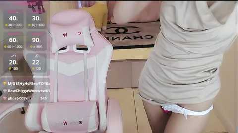 Media: A video featuring a woman in a beige shirt and white panties, sitting on a pink gaming chair, with a virtual chat window overlay.