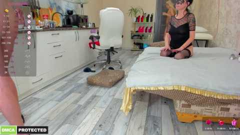 Media: A video shows a woman with fair skin and dark hair, wearing a black lace dress and stockings, kneeling on a bed in a modern, light-colored room with white cabinets, a white chair, and a wooden floor.