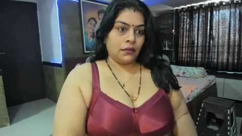 Media: Video of a South Asian woman with medium skin tone and long black hair, wearing a red satin bra, standing in a modern bedroom with a bed, curtains, and a framed picture on the wall.