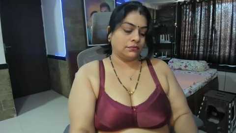 Media: Video of a South Asian woman with medium brown skin and dark hair, wearing a burgundy bra and gold necklace, sitting in a dimly lit bedroom with floral-patterned bedding and brown curtains.