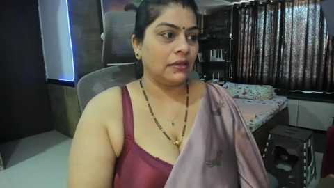 Media: A video shows a middle-aged South Asian woman with medium skin tone, dark hair, and a serious expression. She wears a maroon sleeveless top and a pink shawl. The room has a bed, curtains, and a wooden cabinet in the background.