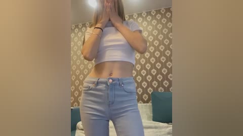 Media: Video of a young woman in a white crop top and light blue jeans, hands covering her face, standing in a bedroom with patterned wallpaper and a bed.