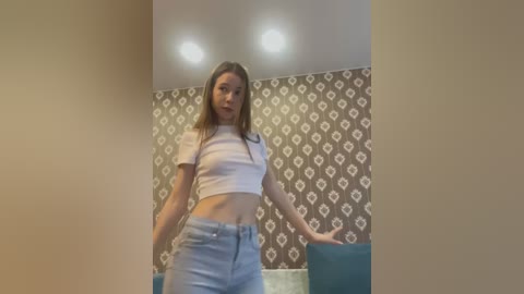 Media: Video of a young woman with straight brown hair, wearing a white crop top and light blue jeans, standing in a room with brown and white patterned wallpaper and teal furniture.