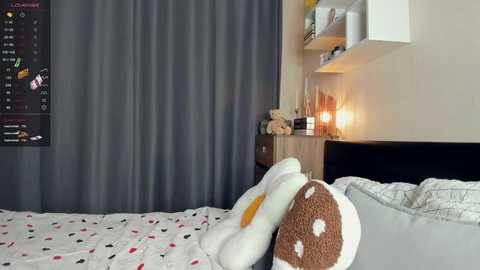 Media: Video of a modern bedroom with a white bed adorned with a plush, brown and white teddy bear and a pillowcase featuring colorful polka dots. Dark gray curtains and a white shelf with a lamp and books are visible.