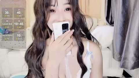 Media: Video of a young Asian woman with long, wavy black hair, wearing white lingerie, smiling while holding a phone to her mouth, in a cozy, dimly lit room with clothes and a white leather couch in the background.