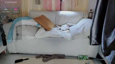 Media: Video of a disheveled white couch with messy white and blue blankets, a brown pillow, and scattered clothes on the floor in a dimly lit room.