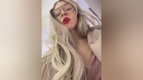 Video of a blonde woman with long, wavy hair, wearing red lipstick, glasses, and a beige jacket, posing in a bright, indoor setting.