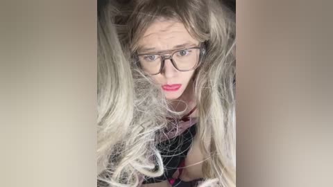 Media: Video of a middle-aged woman with long, messy, platinum blonde hair, wearing black-rimmed glasses and red lipstick. She appears distressed, looking down.