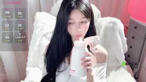 Media: Video of a young East Asian woman with long black hair, drinking from a milk bottle, in a cozy, pink-walled room.