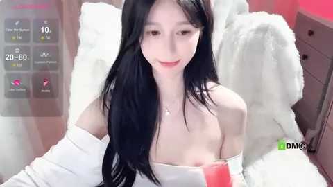 Media: A video of an East Asian woman with long black hair and fair skin, wearing a white off-shoulder top, sitting on a white fur blanket, holding a pink drink.