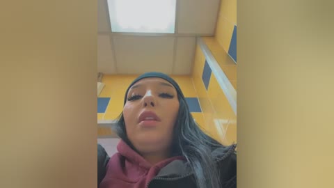 Media: Video of a young Asian woman with long black hair, wearing a gray headscarf and a dark jacket, standing in a yellow, fluorescent-lit hallway with rectangular windows.