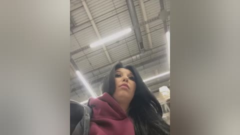 Media: A video captures a woman with long black hair, wearing a maroon hoodie, standing in an industrial warehouse with exposed pipes and a high ceiling.