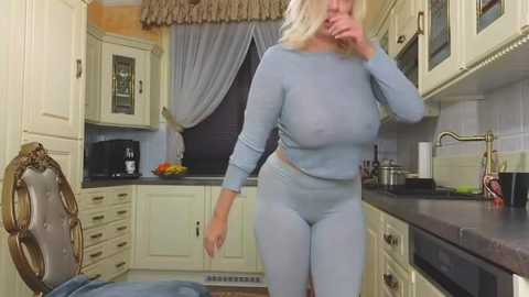 Media: Video of a blonde woman in a light blue, long-sleeve crop top and matching leggings, standing in a modern kitchen with cream cabinets and a black countertop, holding her face with her hand.