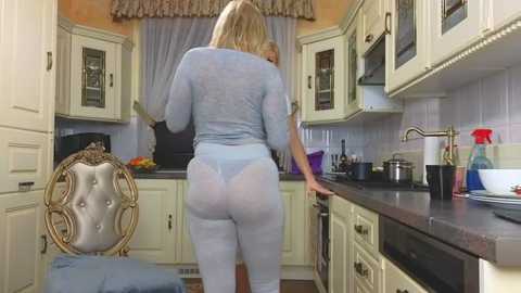 Media: Video of a blonde woman in light blue lingerie, back view, cooking in a vintage-style kitchen with cream cabinets, floral curtains, and a wicker chair.