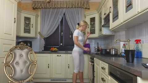 Media: Video of a blonde woman in a white top and pants cooking on a kitchen counter with white cabinets and a black sink.