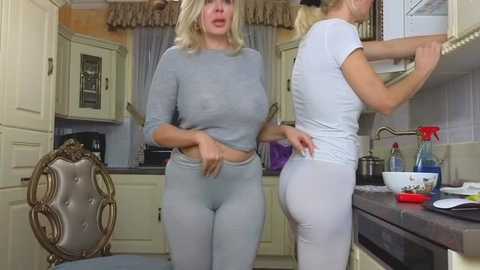 Media: Video of two women in tight, light gray workout outfits, one with a curvy physique and large breasts, the other with a slender frame, in a modern kitchen with beige cabinets and stainless steel appliances.