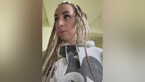 Media: Video of a woman with fair skin and long, blonde dreadlocks wearing a grey hoodie with large, abstract geometric patterns. She stands indoors, facing slightly to the side, with a neutral expression.