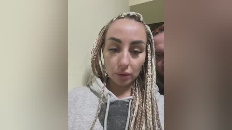 Media: Video of a young woman with fair skin, wearing braided hair extensions and a grey hoodie, leaning against a pale yellow wall.