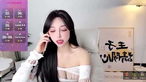 Media: A video of an East Asian woman with long black hair, fair skin, and a slender build, wearing a white off-shoulder sweater, talking on her phone. Background features a purple digital screen, white walls, and a framed artwork.