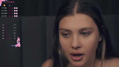 Media: A close-up video of a young, light-skinned woman with long, dark brown hair, wearing a pink top, in a dimly lit room. Her expression is intense and she appears to be speaking.