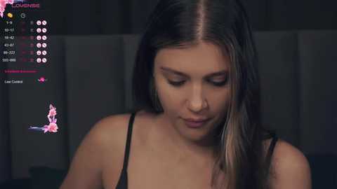 Media: Video of a young woman with straight brown hair and light skin, wearing a black tank top, looking down with a serene expression. Background features dimly lit curtains and digital video game elements on screen.