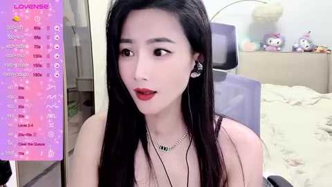 Media: A video of an Asian woman with long black hair and red lipstick, wearing a black top and silver necklace, in a bedroom with plush toys, bed, and a purple chair.