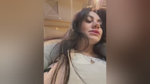 Media: Video of a woman with long dark hair, wearing a beige jacket and white shirt, lying on a bed with a beige blanket, in a dimly lit room with a vent on the ceiling.