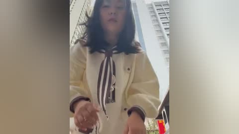 Media: A low-angle video captures an Asian woman in a white blazer and black-and-white striped scarf, holding a small object in a narrow, urban setting with tall buildings and a red canister visible in the background.