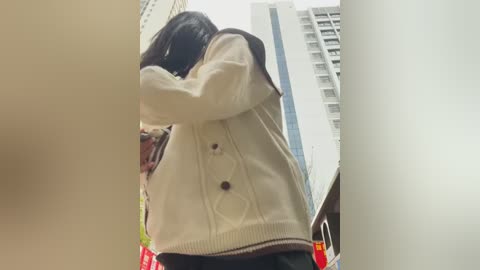 Media: Video of a young person with long black hair, wearing a white sweater and dark skirt, hugging a person out of frame, set against a cityscape with tall buildings.