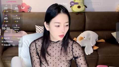 Media: Video of an Asian woman with long black hair, wearing a sheer black polka-dot top, sitting on a brown leather couch. Background includes plush toys and a light-colored blanket.