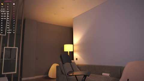 Media: A modern, dimly-lit hotel room with minimalist decor features a large white wall, a yellow lamp, two black chairs, a beige ottoman, and a vertical clock displaying city lights.