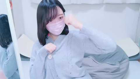 Media: Video of an East Asian woman with straight black hair, pale skin, and a light pink face mask, wearing a light grey cardigan and jeans, sitting on a white bed, indoors.