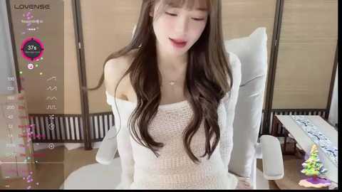 Media: A video of an East Asian woman with long, wavy brown hair, fair skin, and red lipstick, wearing a white off-shoulder knit sweater. She sits on a white chair in a modern, minimalist room.