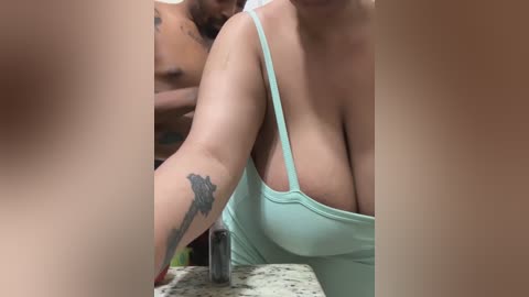 Media: Video of a topless, light-skinned woman with large breasts, wearing a turquoise tank top, exposing cleavage, leaning over a bathroom sink with a tattoo on her upper arm.