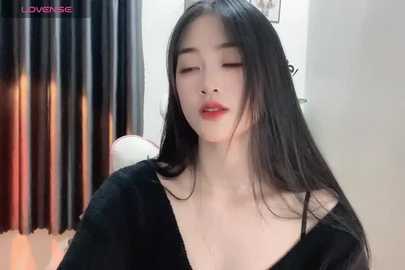 Media: Video of an Asian woman with long black hair, fair skin, wearing a black off-shoulder top, standing indoors with blurred curtains in the background.