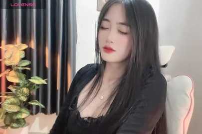 Media: Video of a fair-skinned Asian woman with long black hair, wearing a black lace top, seated indoors, with a floral arrangement and curtains in the background.