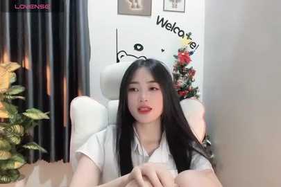 Media: Video of a young Asian woman with long black hair and fair skin, wearing a white shirt, seated in a white chair in a modern room with black curtains, a Christmas tree, and \"Welcome\" sign on the wall.