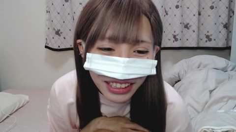 Media: Video of an Asian woman with straight, shoulder-length brown hair, wearing a white surgical mask, kneeling on a bed with white sheets, smiling, with a man's hand visible at the bottom.