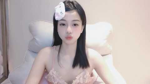 Media: Video of an East Asian woman with straight black hair, a white flower, and pink lace lingerie, sitting on a white cushion, in a soft, pastel-toned room.