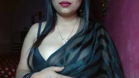 Media: Video of a woman with long black hair, medium skin tone, wearing a sheer black shawl covering a black bra, red lipstick, and a silver necklace. Background features a pink wall and a cluttered room.