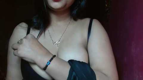 Media: Video of a woman with light brown skin and medium build, wearing a black bra and necklace, with a black bracelet on her wrist, against a dark, textured wall.