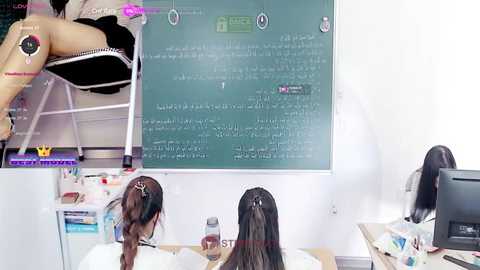 Media: Video of two women studying in a classroom, one sitting on a stool, the other facing a greenboard with Arabic text. White walls, cluttered desk, and a computer screen visible.