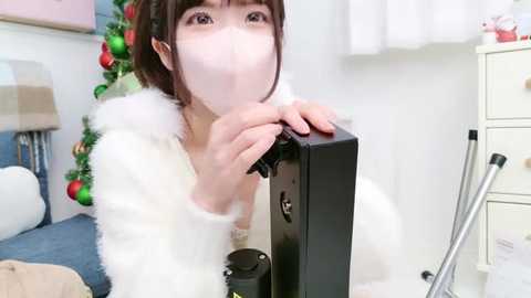 Media: A video of an Asian woman in a white fur-trimmed coat, wearing a face mask, holding a black microphone in a festive room with a decorated Christmas tree and white curtains.